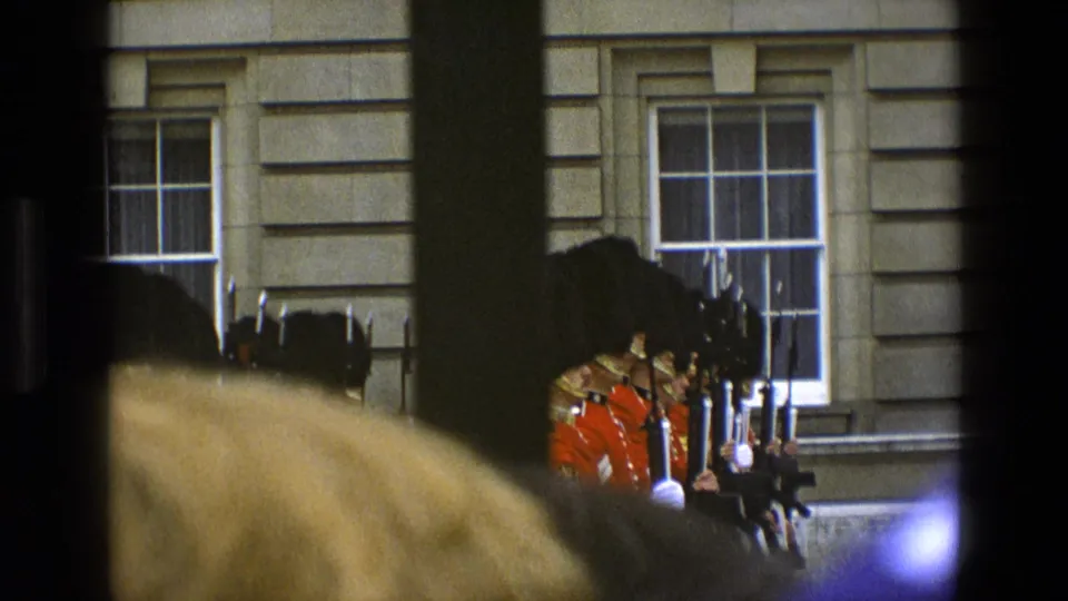 Crowd-obscured 8mm view of guards in red uniforms standing outside a formal stone building in England, with windows behind them.