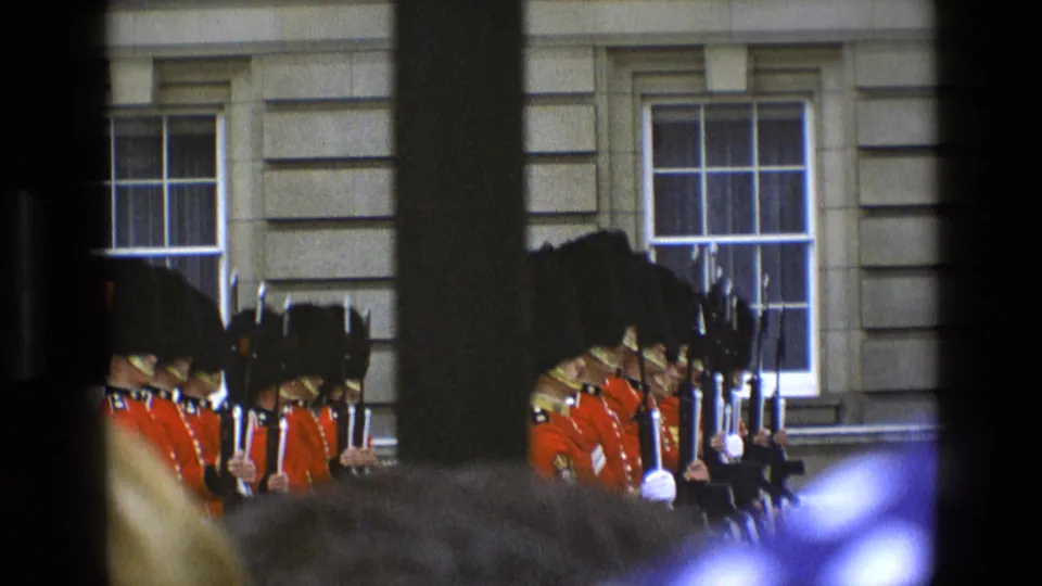Two visible groups of guards in red tunics and black hats stand by a stone palace facade, seen through foreground spectators and framing shadows.