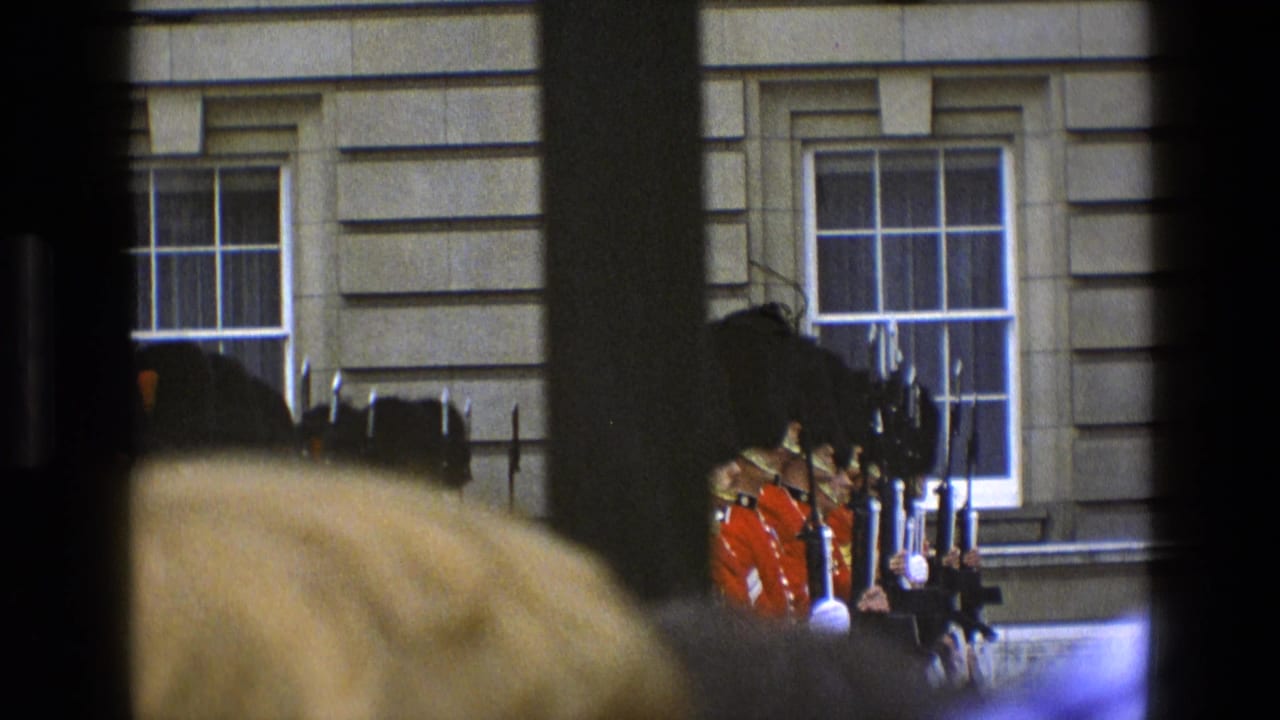 In 1969 England, guards in red uniforms and tall black hats stand before a stone building, partly obscured by a crowd and dark framing.