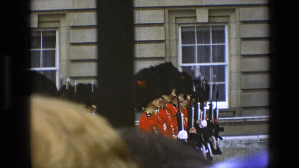 In 1969 England, a crowd-obstructed home-movie view shows ceremonial guards in red uniforms holding rifles before a windowed stone building.