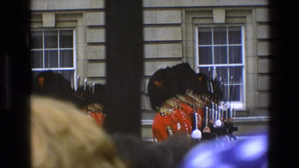 In 1969 England, ceremonial guards in red uniforms and black hats stand with rifles, filmed through a crowd and dark vertical obstructions.