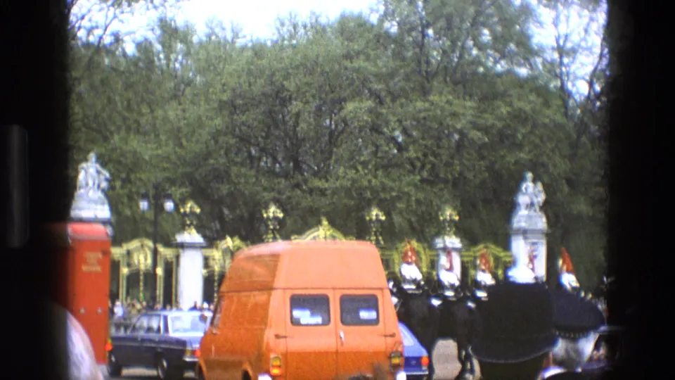 1969 England home-movie frame with a red van, classic cars, ornate gates, and guards marching past spectators.