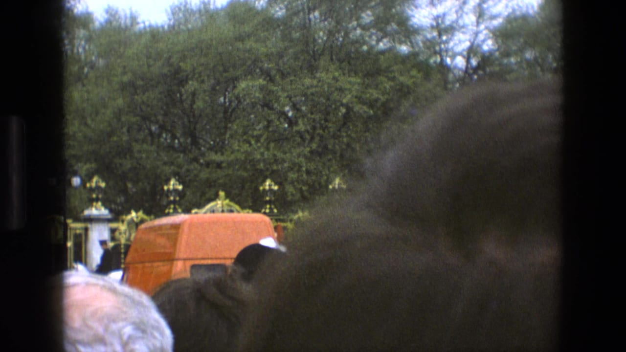 1969 England home-movie frame with crowd heads in the foreground, ceremonial guards by ornate gates, trees, and an orange van.