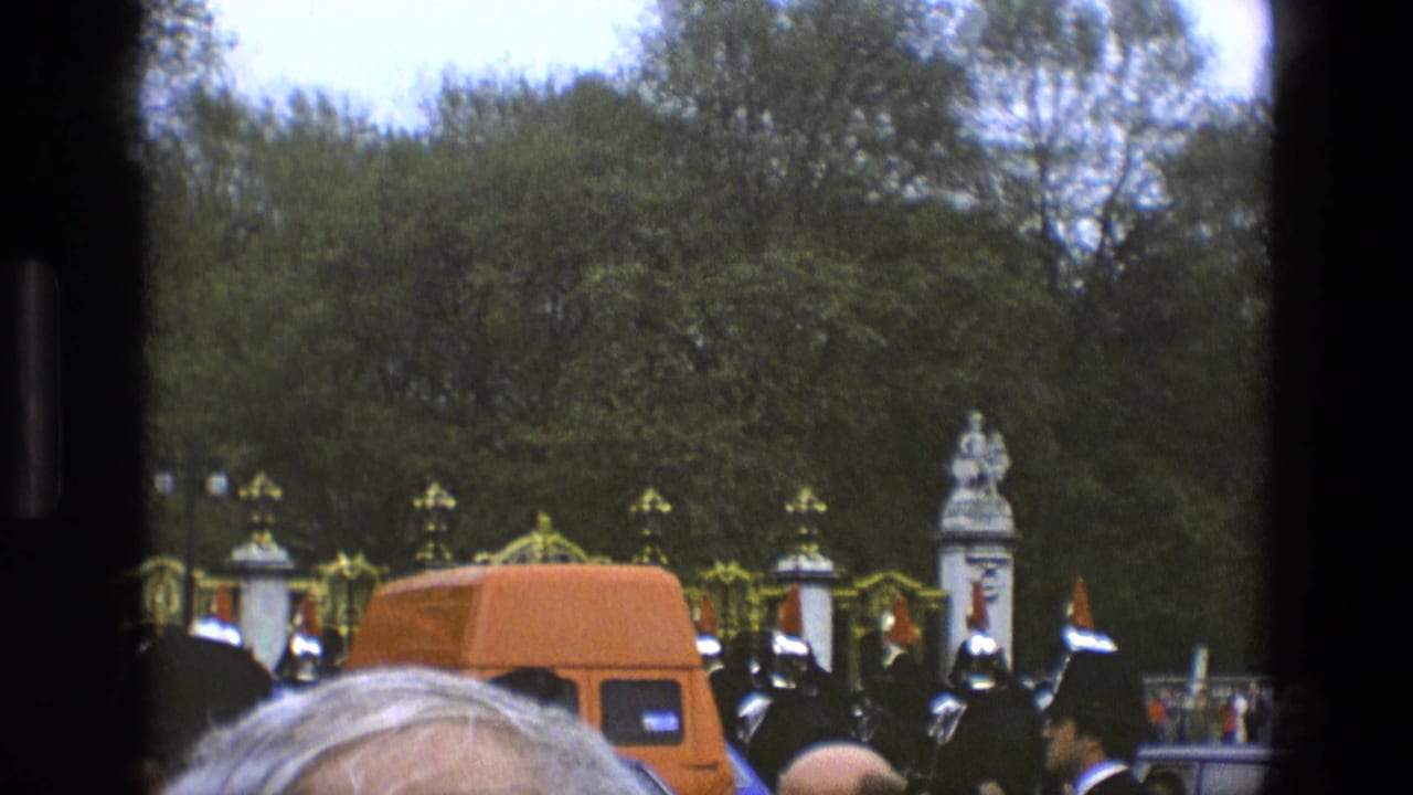 Orange van and mounted ceremonial guards pass in front of ornate gates and trees.