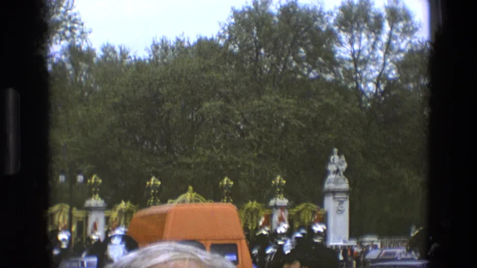 1969 England home-movie view of ceremonial guards, an orange van, vintage cars, crowd heads, and palace-style gates.