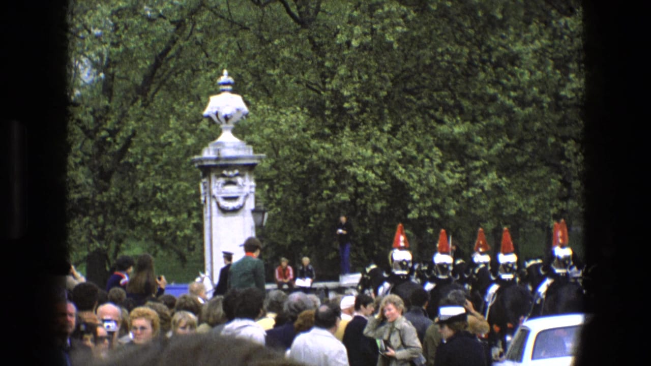 1969 England home-movie frame showing spectators facing mounted ceremonial guards beside a white carved pillar in a leafy setting.