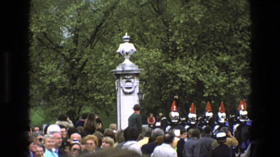 1969 England frame with a dense crowd, mounted ceremonial guards, red headgear, and a white carved pillar in a green outdoor setting.