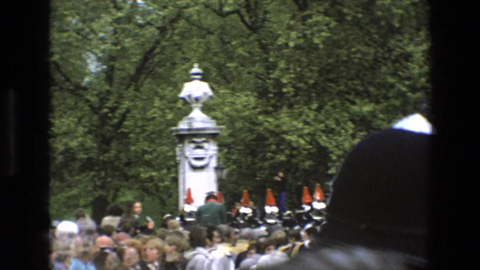 1969 England home-movie frame showing mounted ceremonial guards, spectators, trees, and a white stone pillar, with a dark obstruction at right.