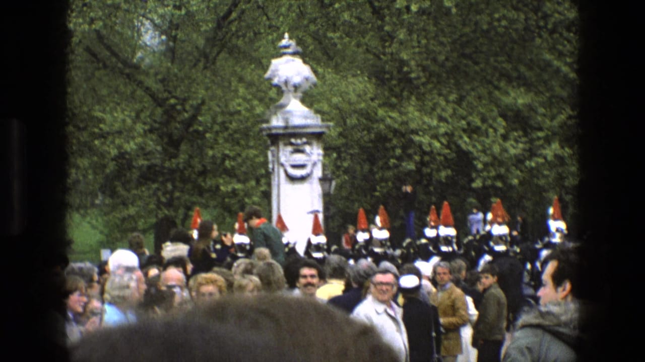 1969 England home-movie frame showing spectators and ceremonial soldiers with red plumes near a white stone monument in a green park.