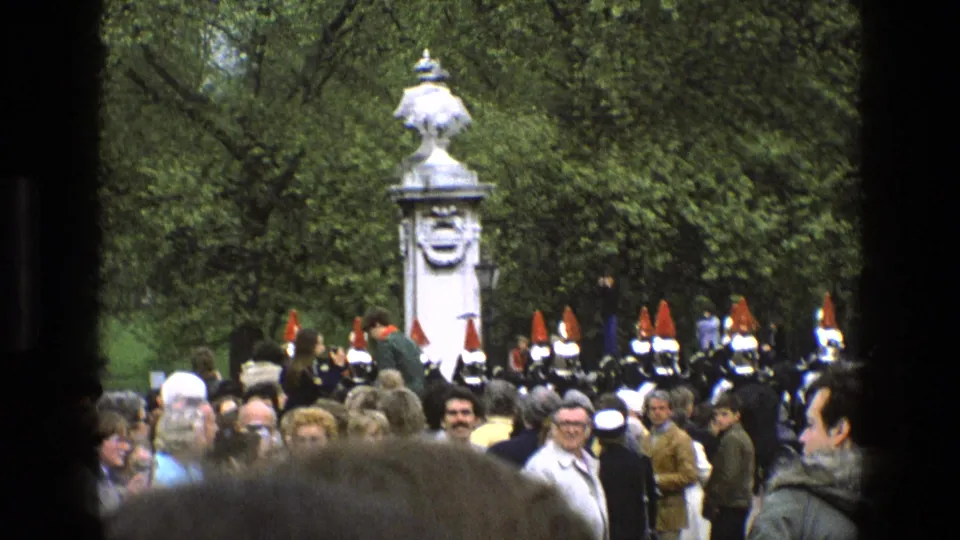 1969 England home-movie frame showing spectators and ceremonial soldiers with red plumes near a white stone monument in a green park.