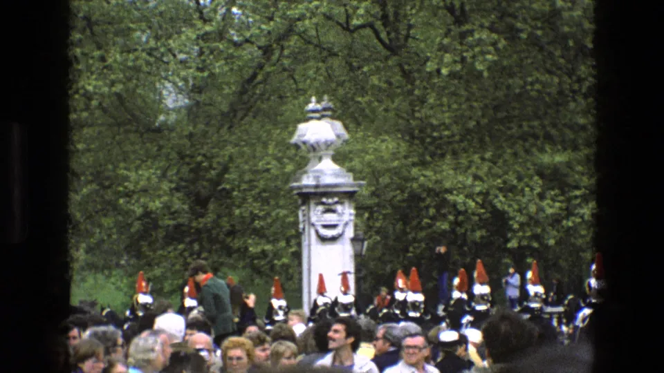 1969 England frame with a foreground crowd, ceremonial soldiers in dark uniforms, red plumes, and a white monument among trees.