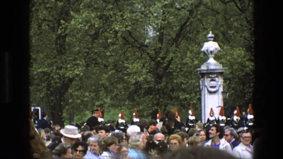 1969 England home-movie view of spectators watching ceremonial soldiers beside a white stone monument and dense trees.
