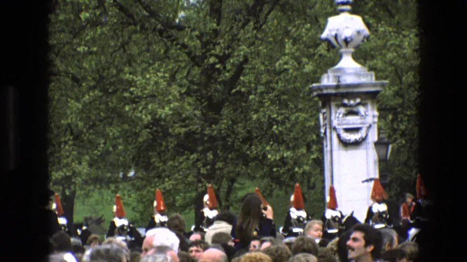 1969 England frame showing a crowded park scene with ceremonial soldiers, red-plumed headgear, trees, and a white stone monument.