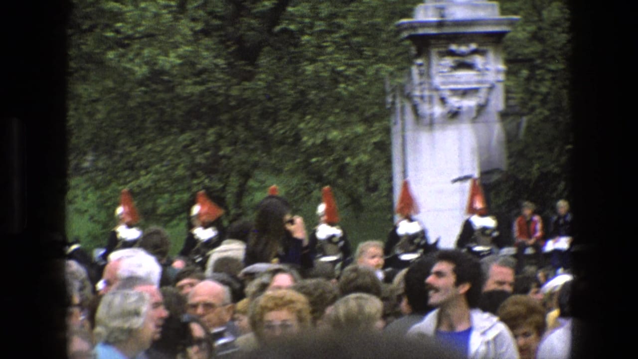 1969 England home-movie frame showing spectators watching ceremonial guards in red tunics near white stone gate pillars and trees.