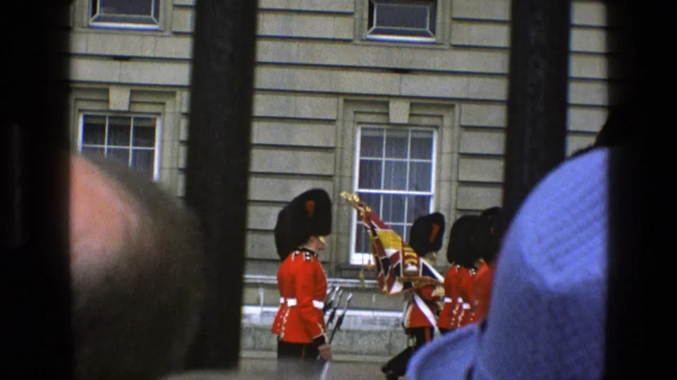1969 England frame showing guards in red tunics and bearskin-style hats marching with flags near a formal stone building.