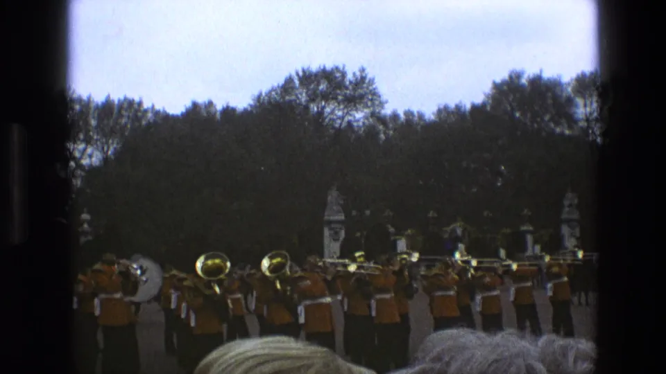 1969 England home-movie frame of a marching military band with brass instruments, seen past spectator heads and a central barrier.
