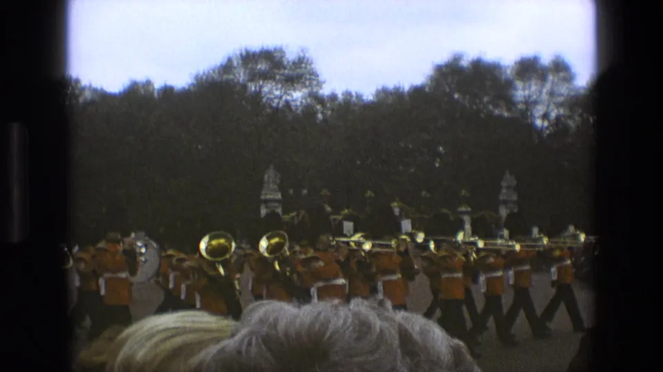 1969 England parade footage showing a uniformed military band marching across the grounds, with spectators and trees in view.