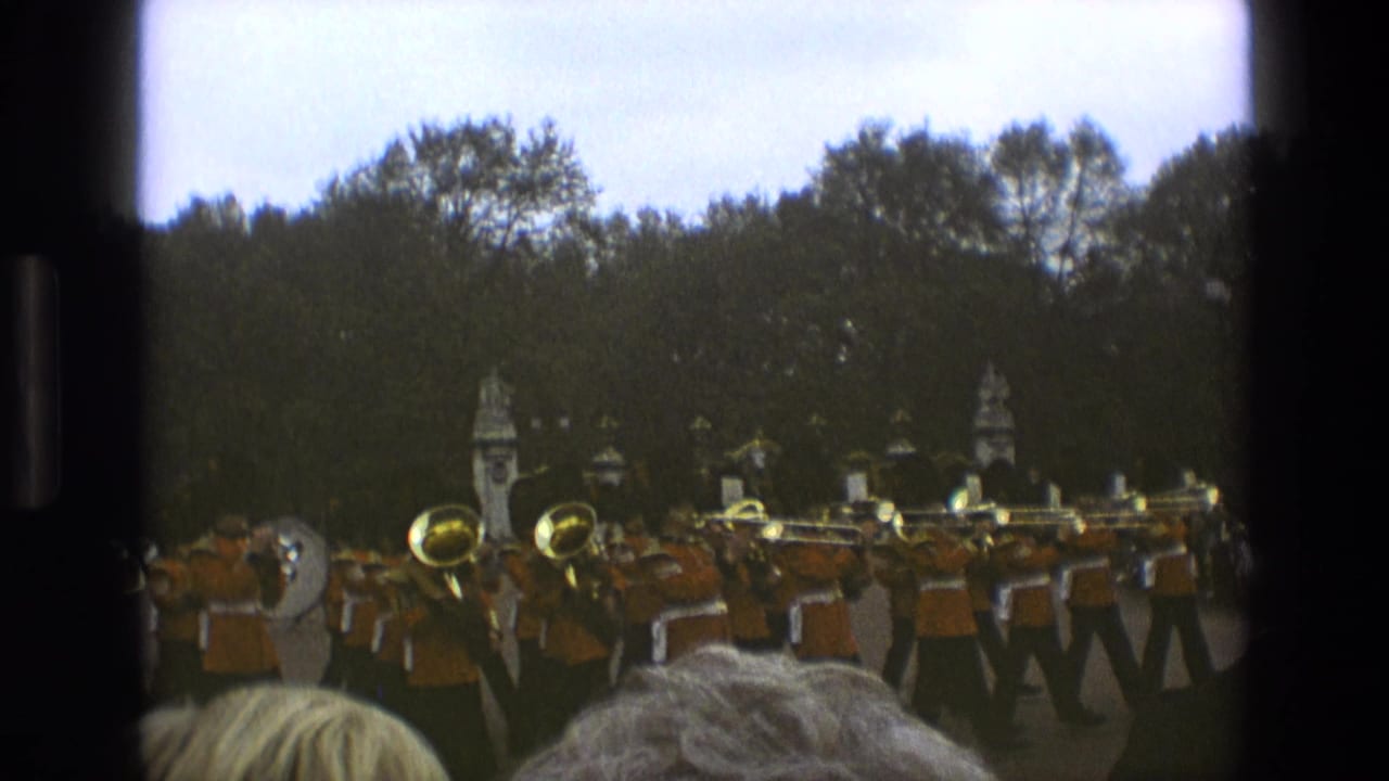 1969 England home-movie frame showing an orange-uniformed marching band performing outdoors behind spectators' heads.