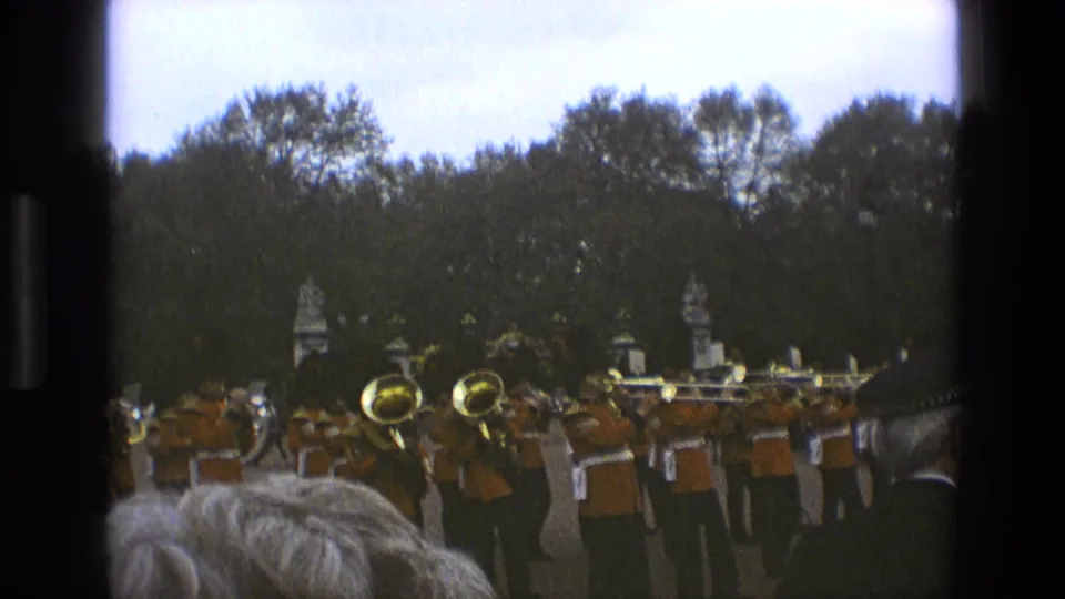 1969 England frame of a brass marching band in orange uniforms seen from behind a crowd at an outdoor event.