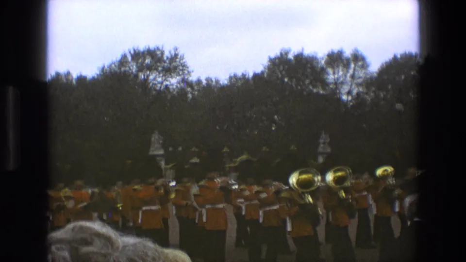 Final 1969 England home-movie frame with a marching band partly obscured by spectators and dark film borders.
