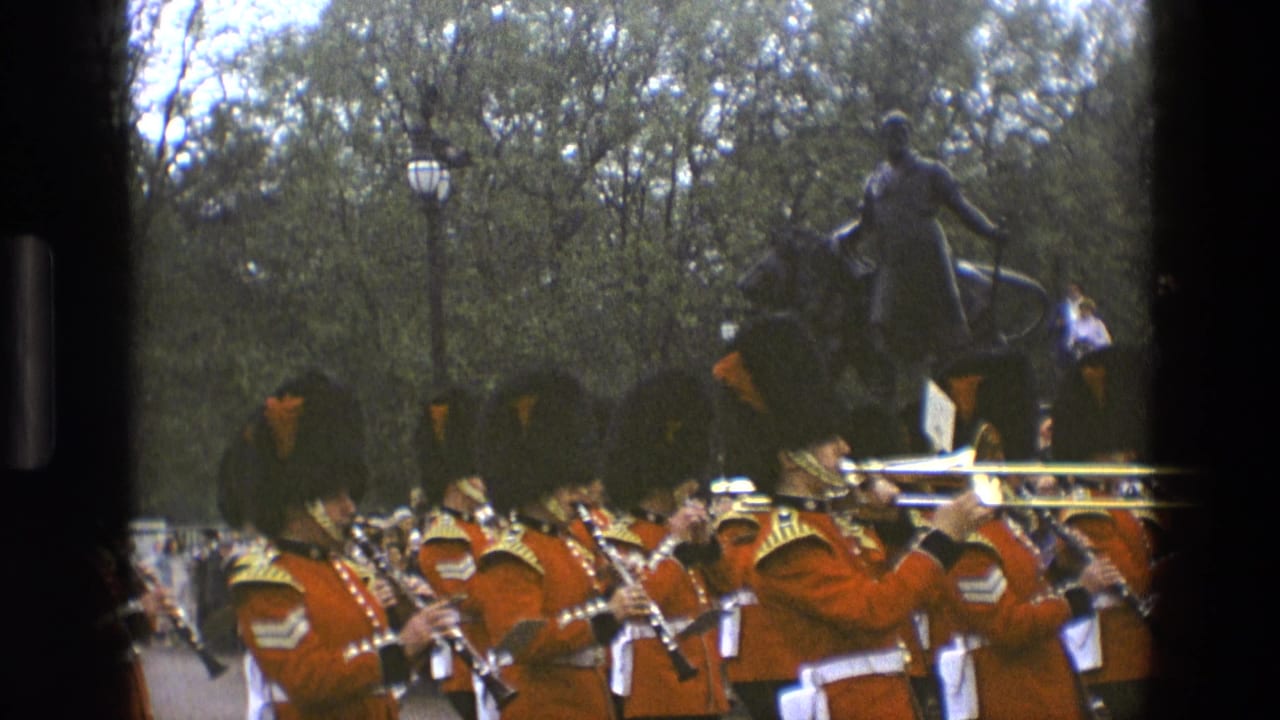 British military band in red uniforms marches past an equestrian statue with spectators behind them.