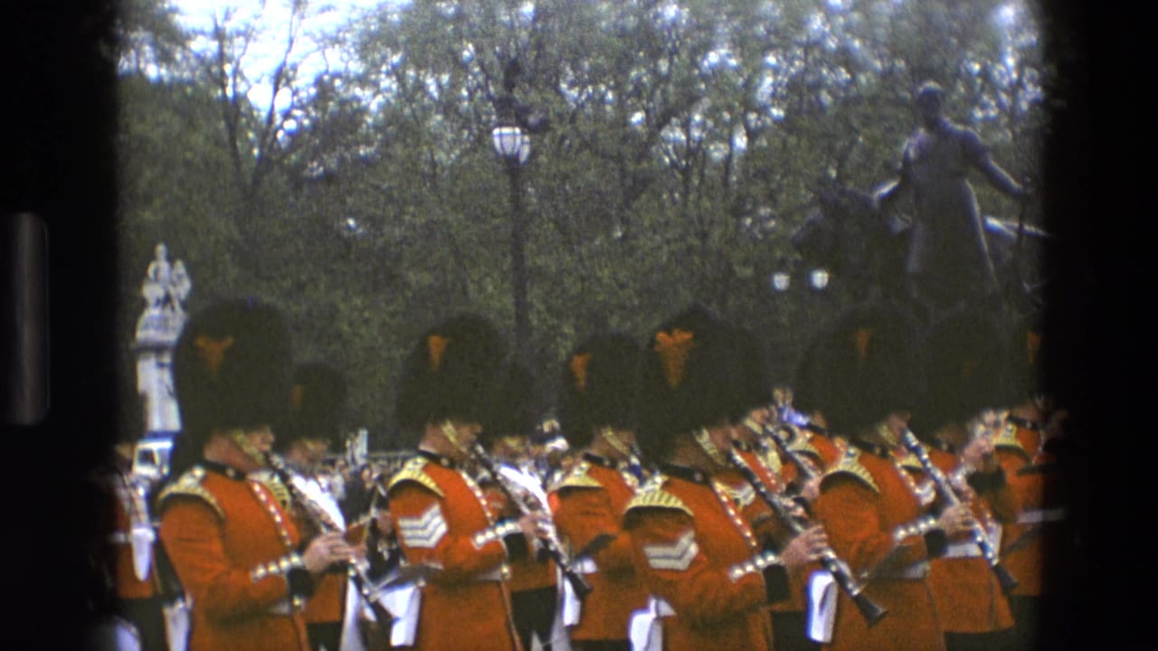 Marching band members in red ceremonial dress play instruments in front of trees and statues.