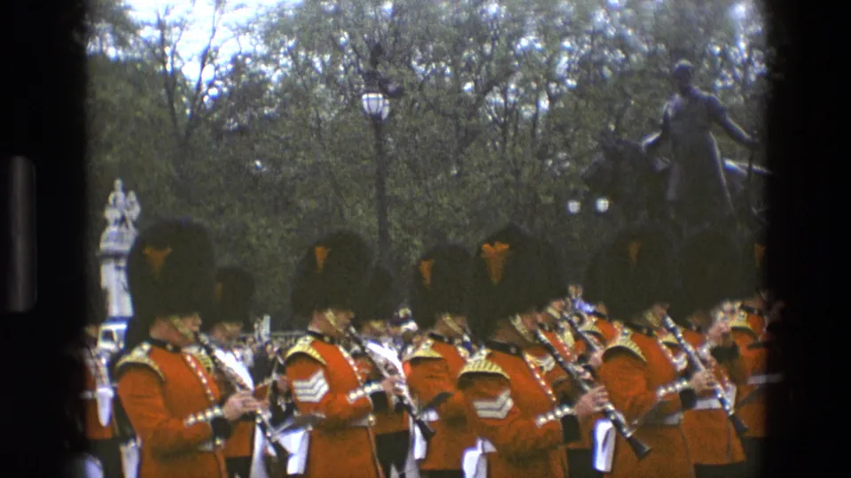 1969 England home-movie frame of uniformed band members playing as they march through a park-like ceremonial area.