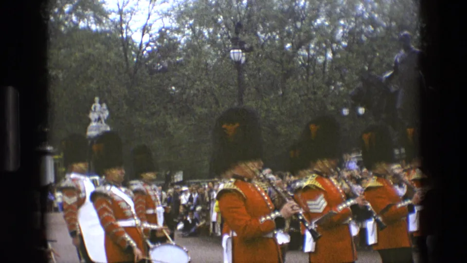 1969 England 8mm frame showing a British ceremonial band moving past crowds, trees, and a distant monument.