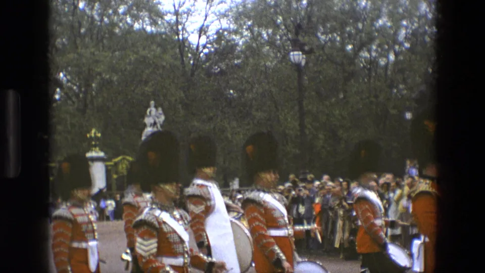 1969 England home-movie frame with red-coated band members, a bass drum, spectators, trees, and a pale monument behind them.