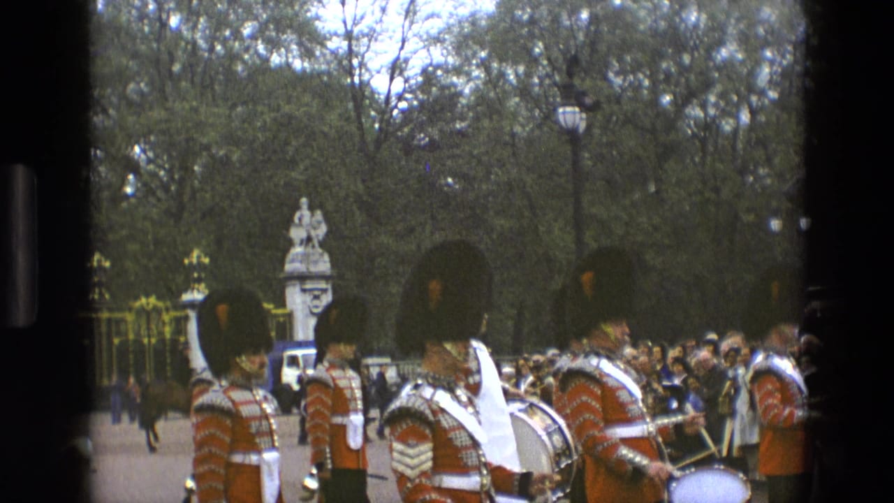 Royal guards in red uniforms march with drums as spectators watch near gates and trees.