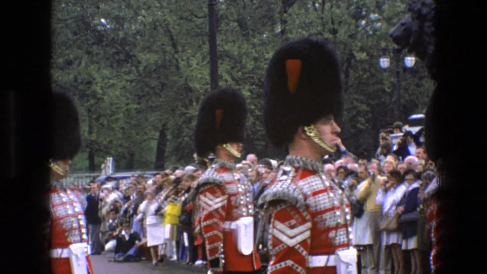 1969 England home-movie frame of royal guards marching past spectators gathered along a tree-lined ceremonial route.