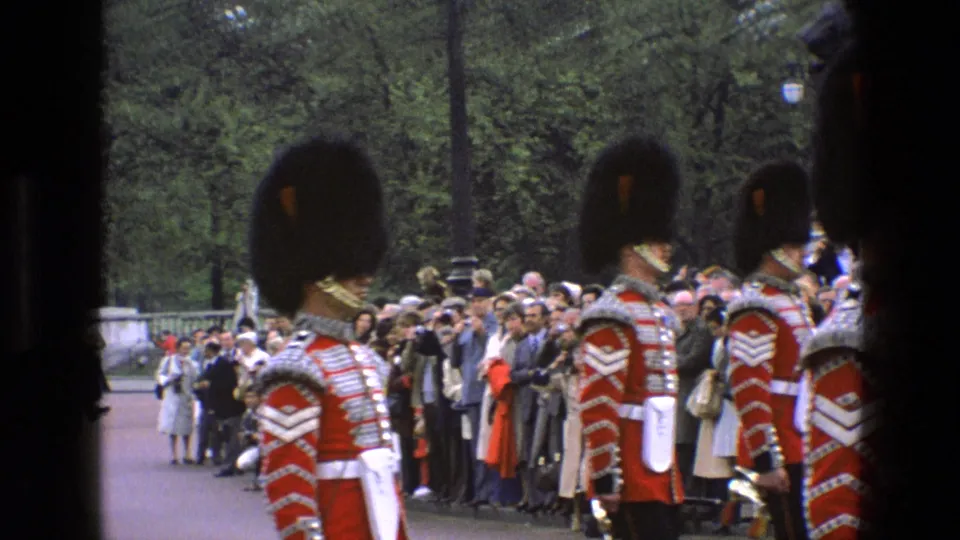 In England in 1969, Grenadier Guards in ceremonial red uniforms and bearskins appear beside spectators outside Buckingham Palace.