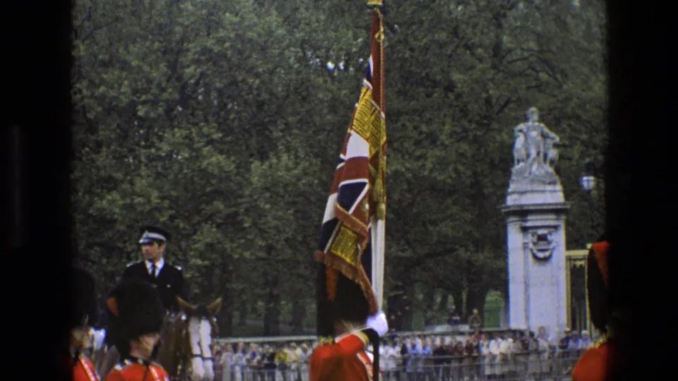 A Union flag and ceremonial guard pass near a mounted officer, spectators, trees, and stone memorial features outside Buckingham Palace.
