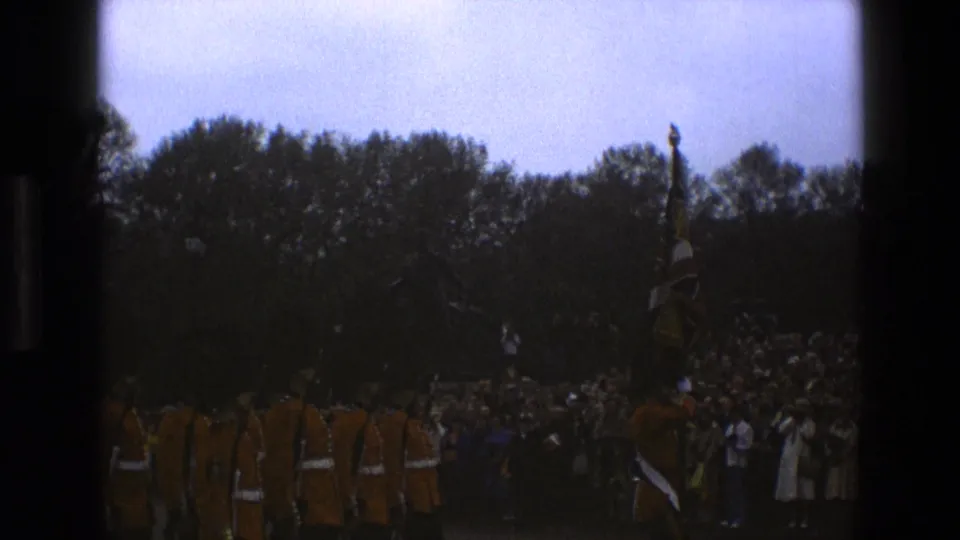 A darker 8mm frame shows guards and a flag moving along the parade route before a dense crowd in England in 1969.