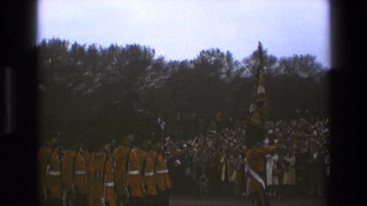 Guards in red uniforms march before a large crowd in an outdoor setting.
