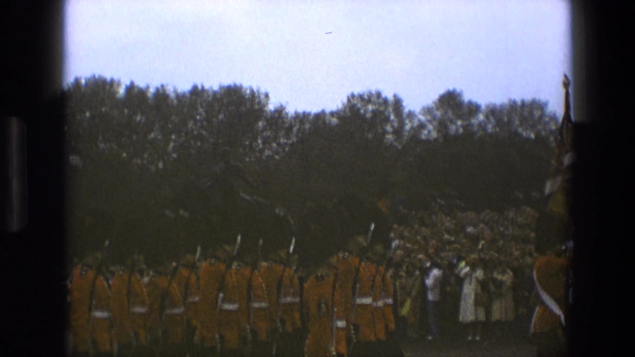 1969 England frame with Queen's Guards in red tunics moving past onlookers in a park-like ceremonial area.