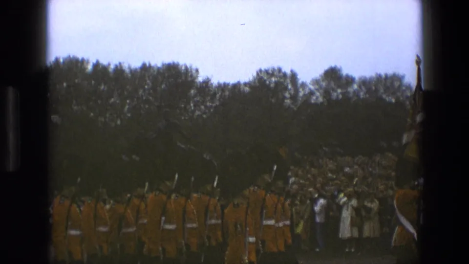 1969 England frame with Queen's Guards in red tunics moving past onlookers in a park-like ceremonial area.