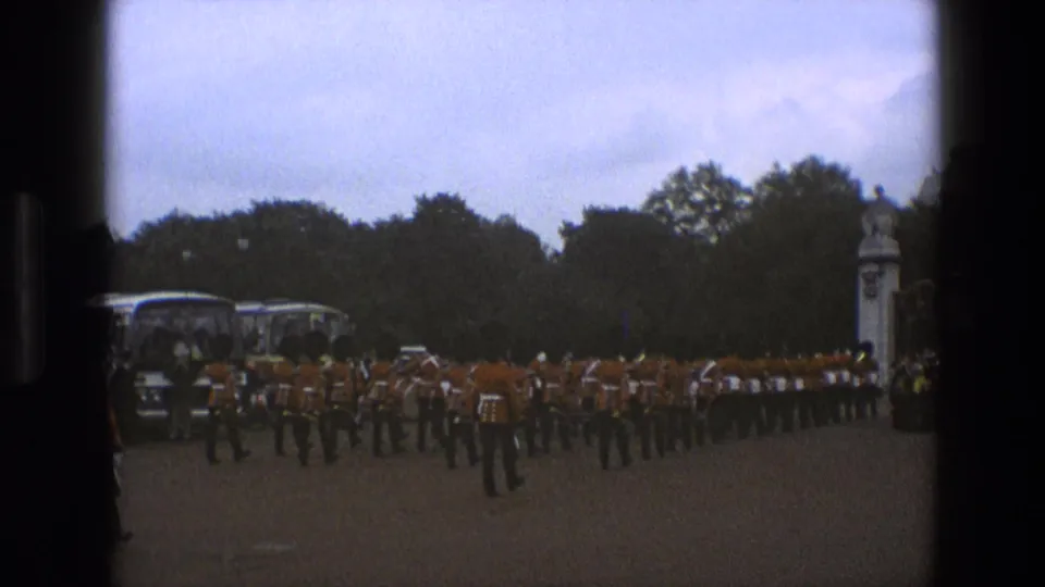 Wide 1969 England frame showing a long line of red-uniformed guards marching near coaches, trees, and a small structure.