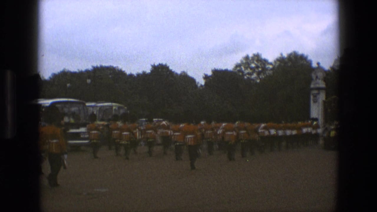 Uniformed marching band in red-orange coats crosses a paved area near trees and buses.