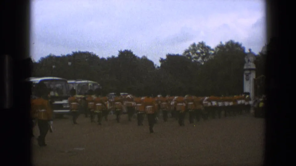 1969 England home-movie frame showing a ceremonial marching formation in red-orange coats and tall black hats near trees and parked buses.