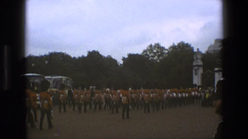 A 1969 England frame shows rows of uniformed marchers in red-orange tunics moving in formation near trees and white gate posts.