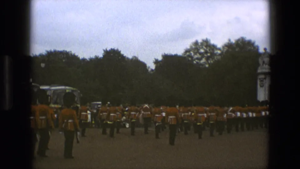 1969 England home-movie frame with a red-orange uniformed formation passing across pavement, with parked buses and trees behind.