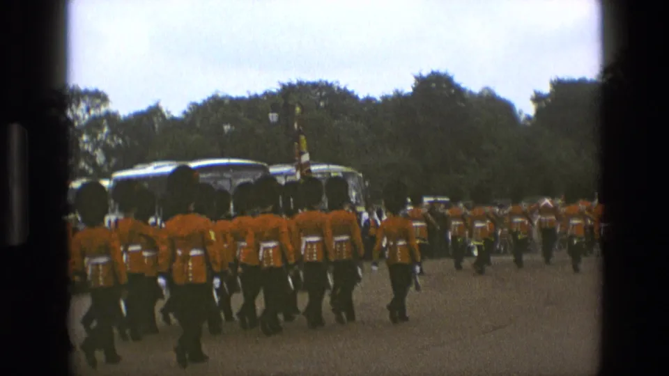 A closer 1969 England frame shows the backs of ceremonial marchers in red-orange coats and tall black hats near parked buses.