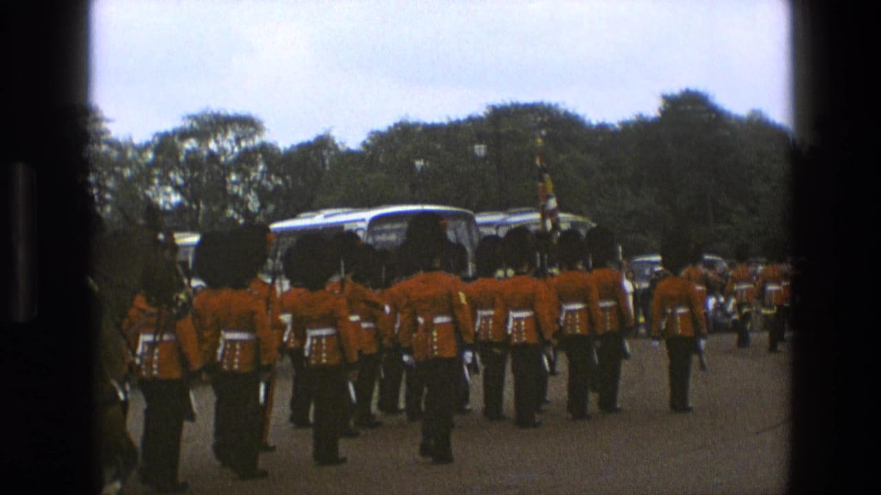 1969 England home-movie frame showing British soldiers in red dress uniforms and tall black hats marching away during a ceremonial parade.