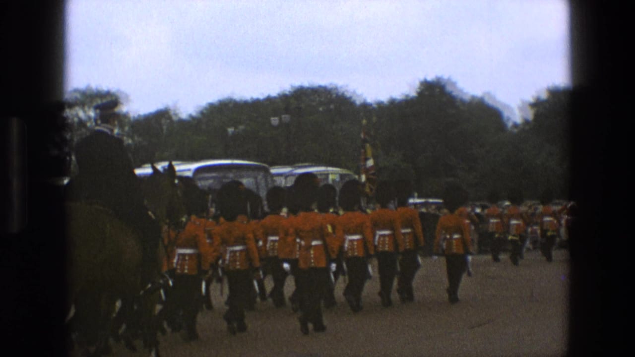 1969 England parade frame with soldiers in red dress uniforms marching behind a mounted officer and ceremonial flag in a public outdoor setting.