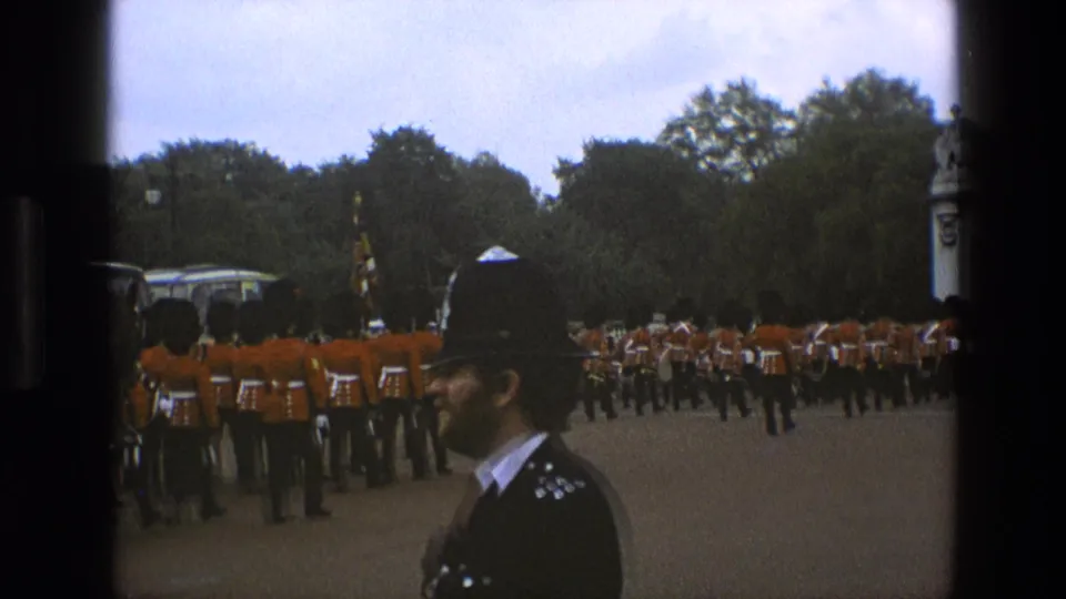 1969 England home-movie frame with a police officer in the foreground and red-uniformed soldiers marching in the background.