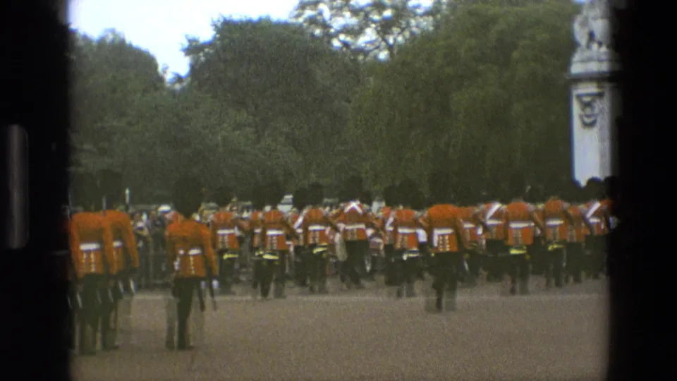 1969 England parade frame showing red-uniformed soldiers gathered along a route, with trees and part of a stone monument visible at the edge.