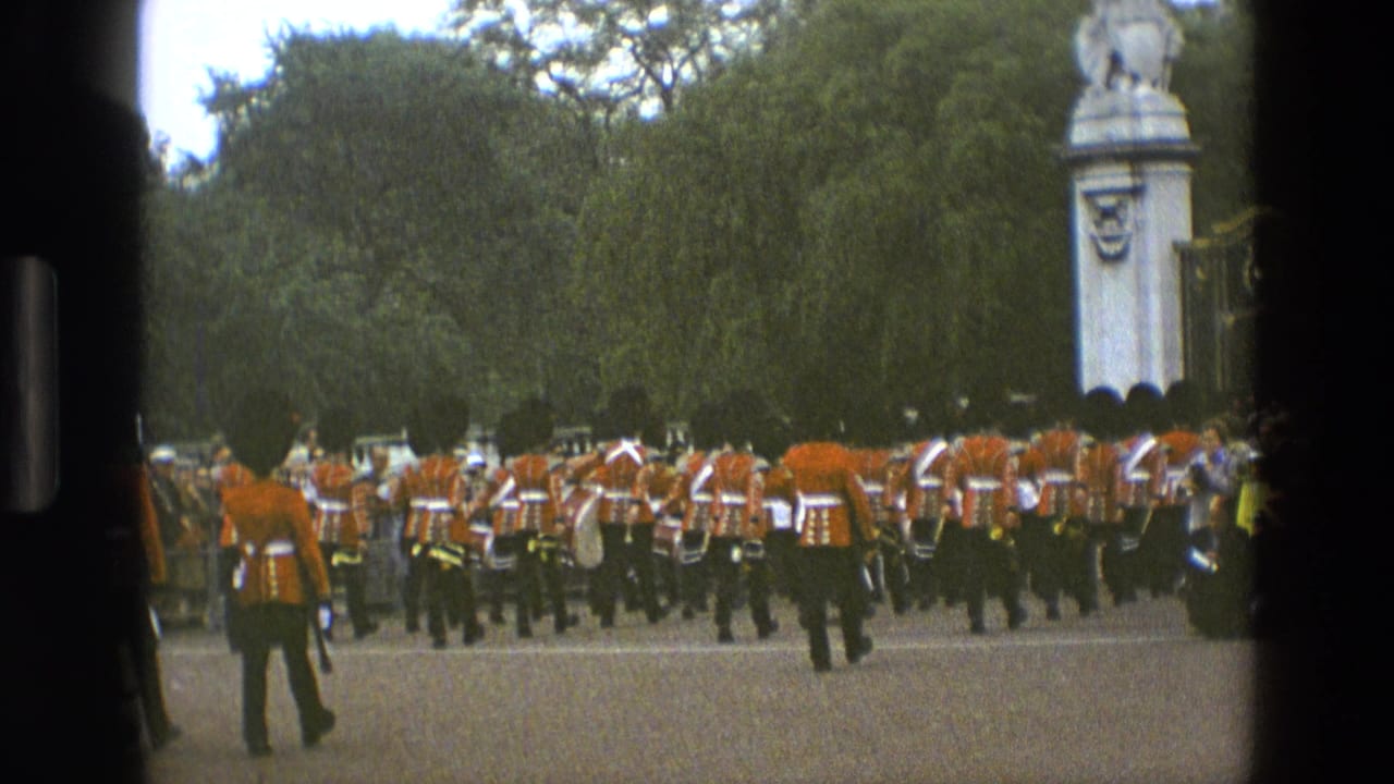 British guards in red tunics march away through a park, with trees and a stone gate structure visible.