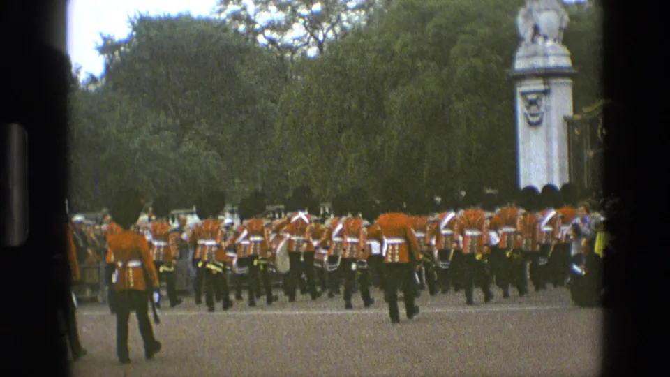 1969 England home-movie frame showing British guards in red tunics and black hats marching away during a ceremony in a tree-lined park.