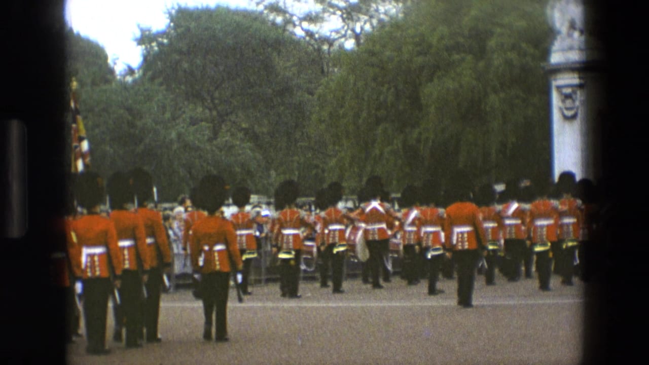 A line of red-coated guards stands or marches near flags, framed by dark film borders and trees.